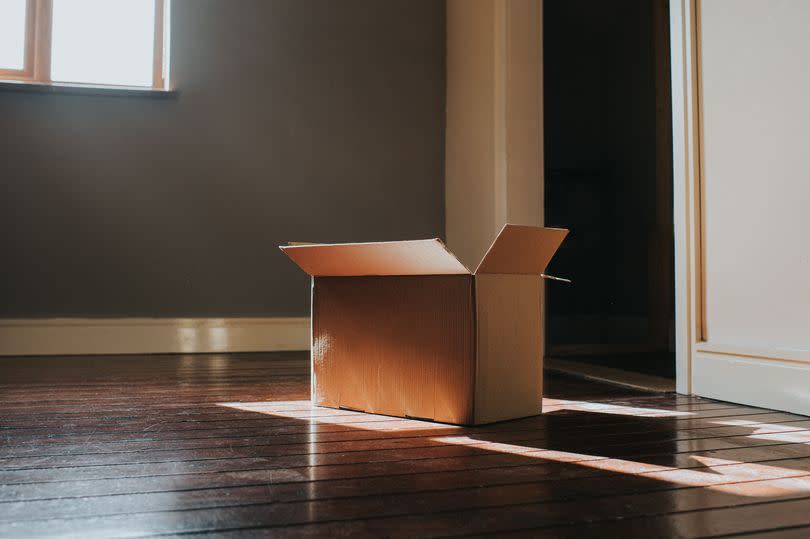 A single, plain, cardboard box sits on a dark wooden floor in a domestic room.