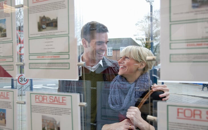 A heterosexual couple are pictured in a warm embrace and smiling as they look into an estate agent window.