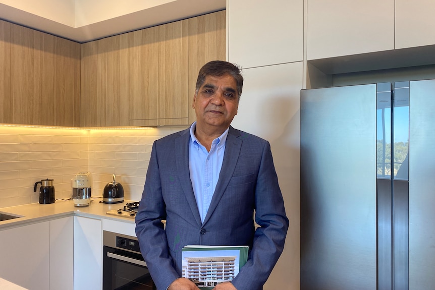 An older Indian man wearing a suit and collared shirt without a tie stands in the kitchen of a modern house.