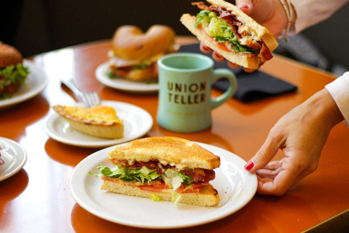 A bacon, lettuce, and tomato sandwich cut crosswise on a white plate. The plate is sitting on an orange table with a seafoam green mug in the background that reads Union Teller. A hand is lifting up the other half of the sandwich. 